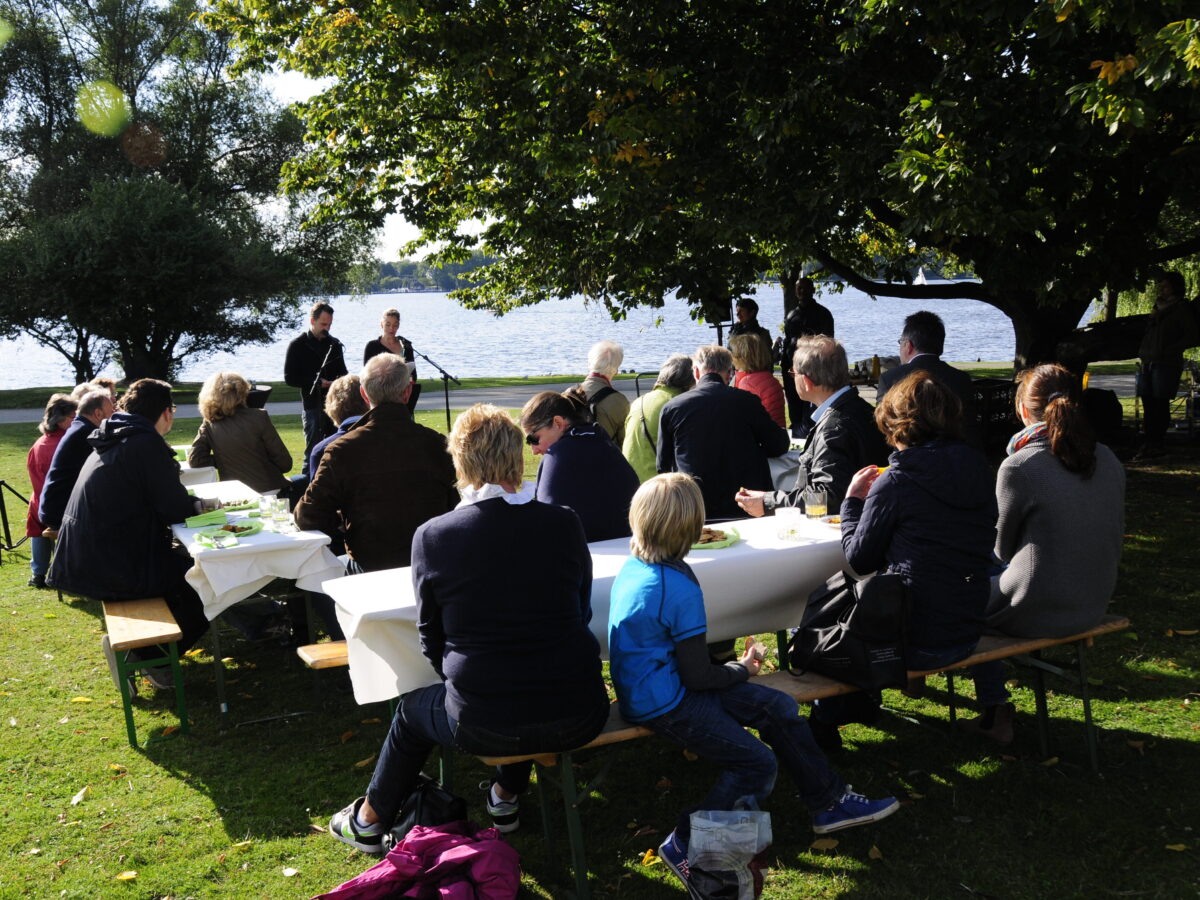 Auf einer Wiese nahe am Wasser stehen Tische und Bierbänke, auf denen mehrere Personen sitzen.© Literaturhaus