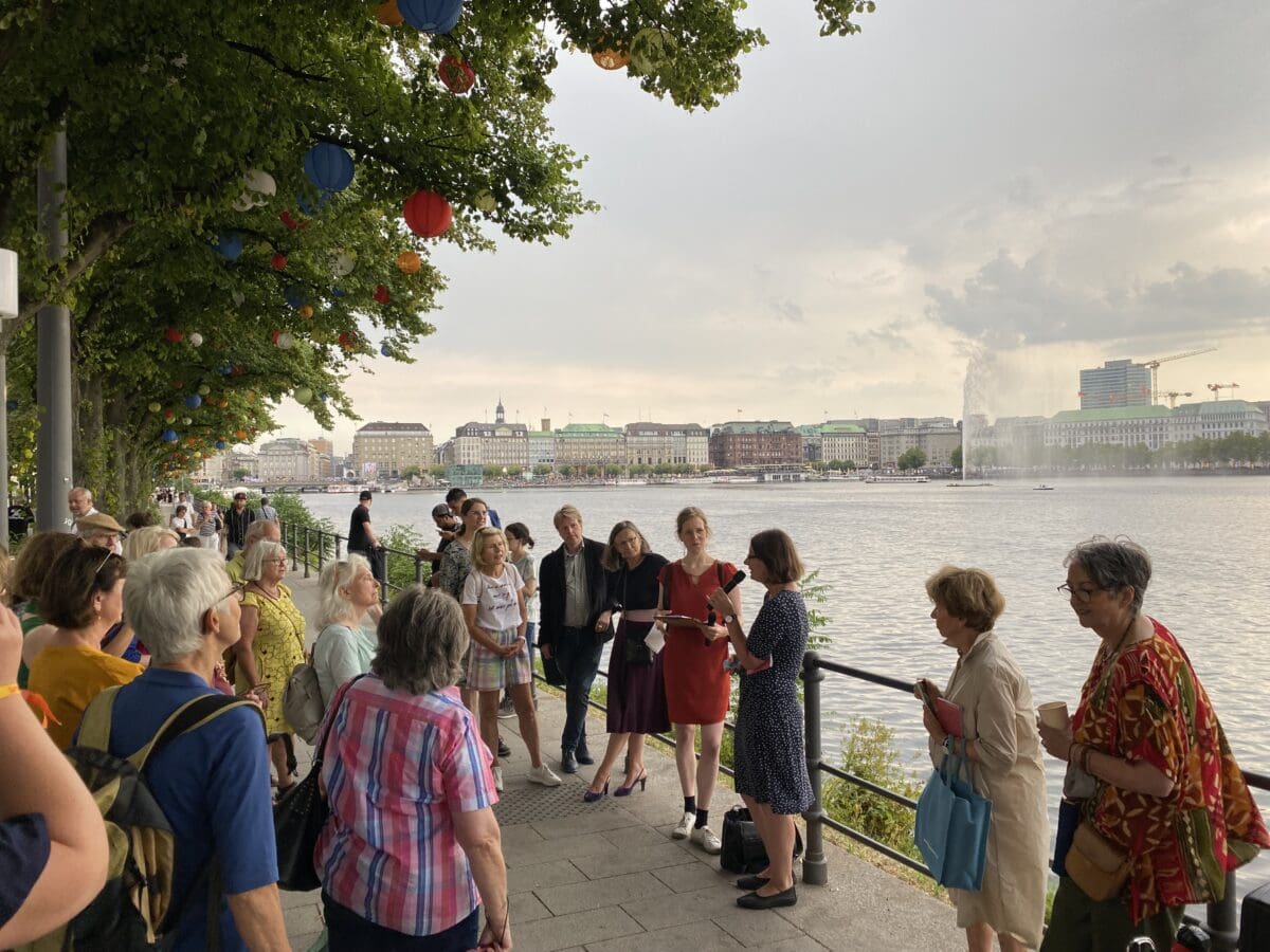 Unter lampionbehängten Bäumen am Rande der Binnenalster stehen mehrere Personen. Eine Frau spricht in ein Mikrofon.© Literaturhaus