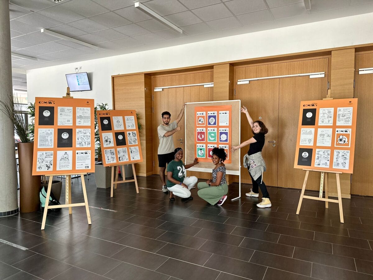 Auf Staffeleien stehen mit den Comixx bedruckte Ausstellungstafeln. Mehrere junge Erwachsene stehen darum herum und präsentieren die Tafeln.