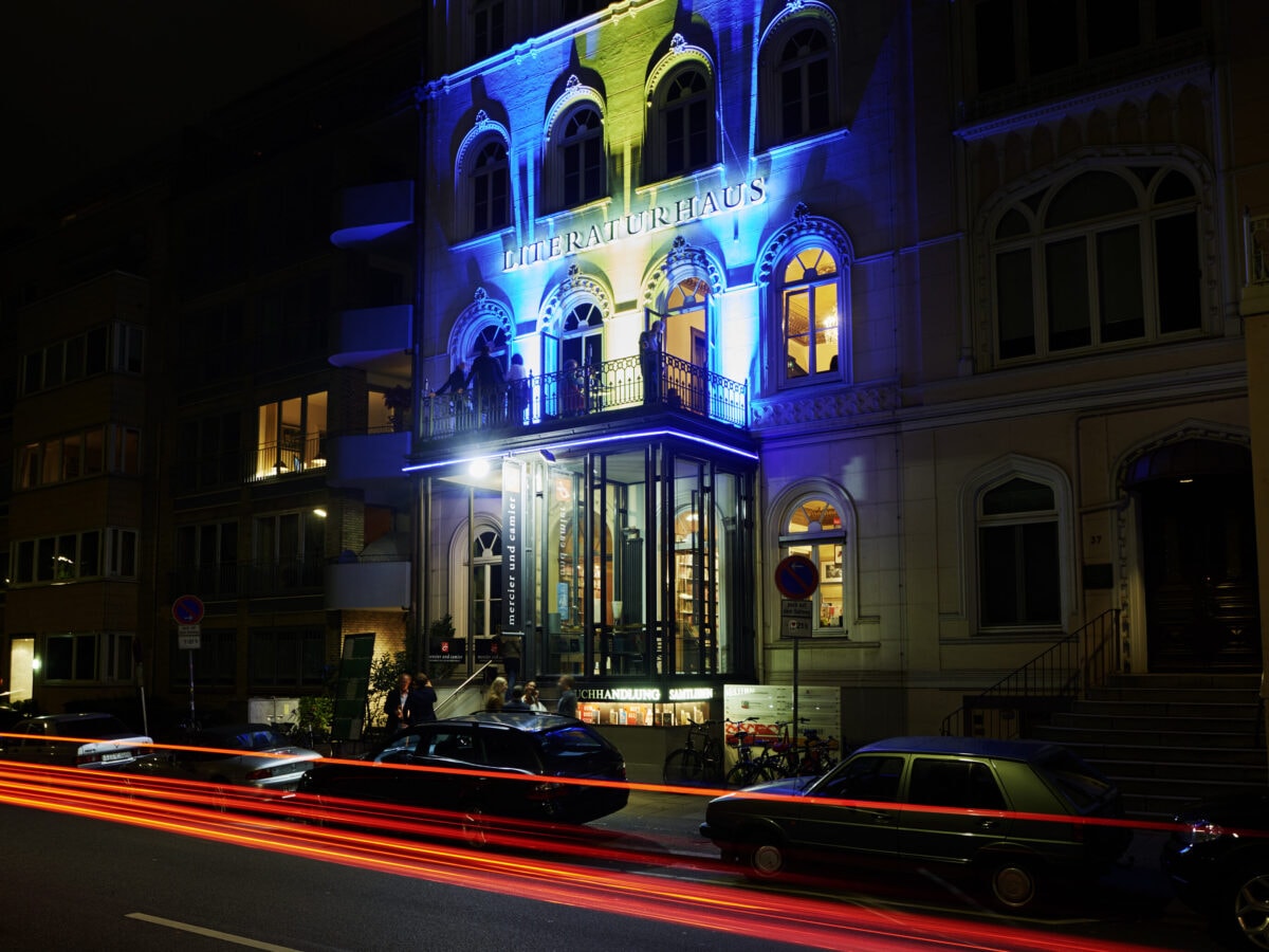 Die Hausfassade bei Nacht und mit Langbelichtung fotografiert. Die roten Rücklichter von vorbeifahrenden Autos sind als Lichtstreifen zu sehen. Die Hausfassade ist blau beleuchtet.© Kathrine Uldbæk Nielsen