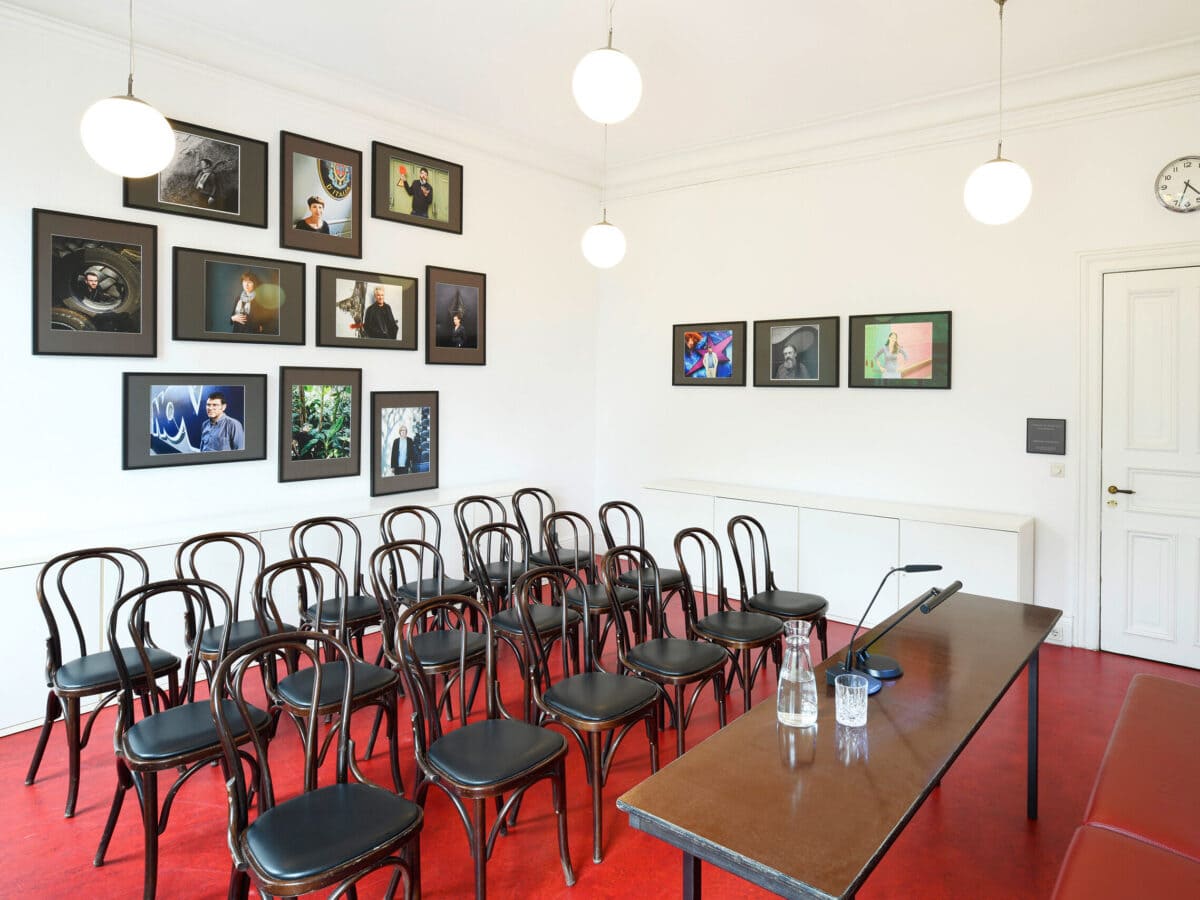 Blick in das Gartenzimmer: Der Fußboden ist rot, an der Wand hängen einige Porträtfotos. Stühle stehen in drei Reihen mit Blickrichtung zu einem Tisch, auf dem ein Mikrofon steht.© Literaturhaus