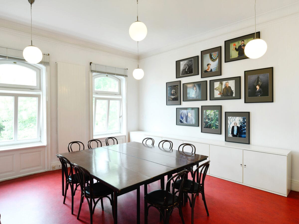 Blick in das Gartenzimmer: Der Fußboden ist rot, an der Wand hängen einige Porträtfotos. In der Mitte steht ein Tisch mit Stühlen drumherum.© Literaturhaus