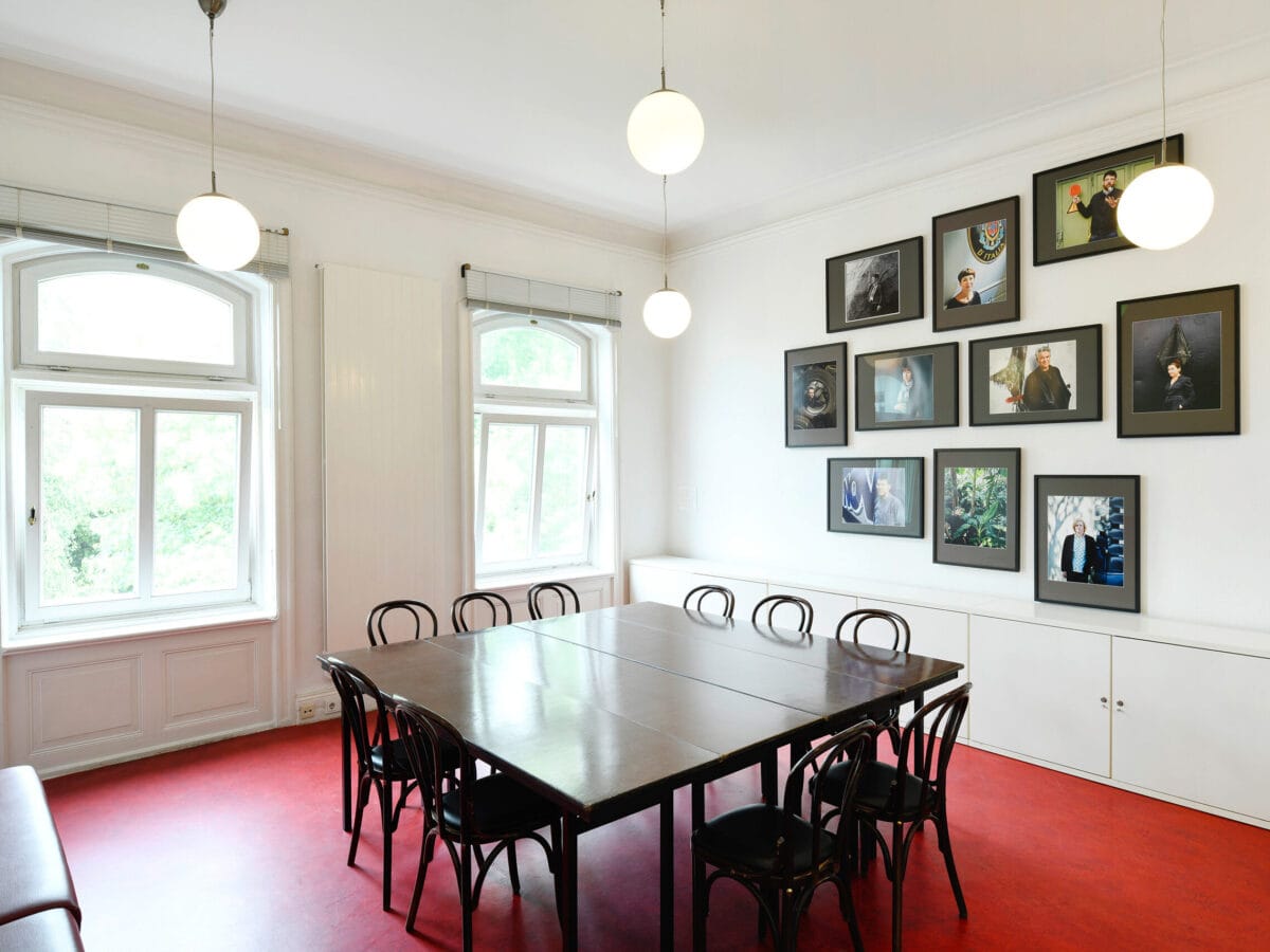 Blick in das Gartenzimmer: Der Fußboden ist rot, an der Wand hängen einige Porträtfotos. In der Mitte steht ein Tisch mit Stühlen drumherum.© Literaturhaus
