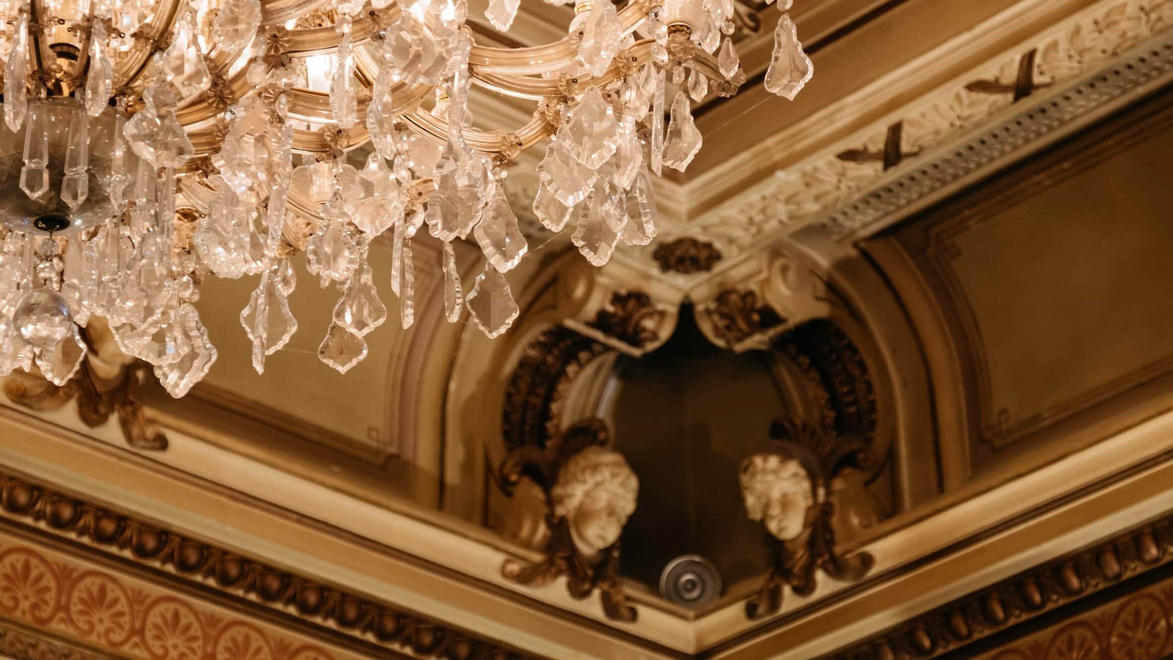 Im Vordergrund: Der Kristallkronleuchter im Eddy-Lübbert-Saal. Im Hintergrund: Zwei Engelköpfe in Stuck eingefasst in einer Ecke des Saals.© Literaturhaus