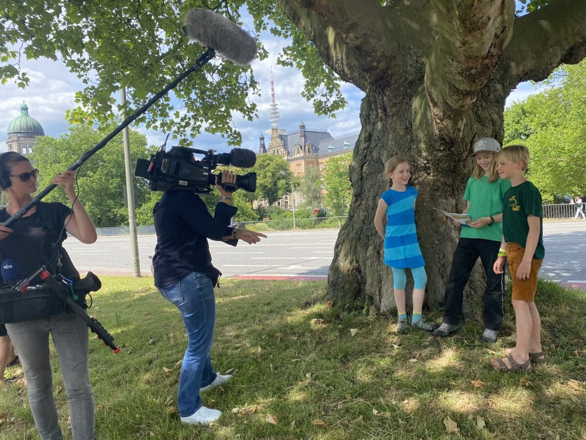 Drei Kinder stehen vor einem Baum. Eines der Kinder hält einen Rallyebogen für eine Stadtsafari in den Händen.© Literaturhaus