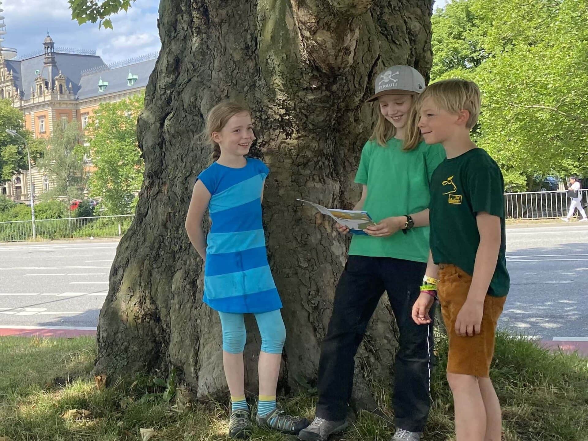 Drei Kinder stehen vor einem Baum. In der Hand halten Sie einen Flyer für die Stadtsafari.© Literaturhaus